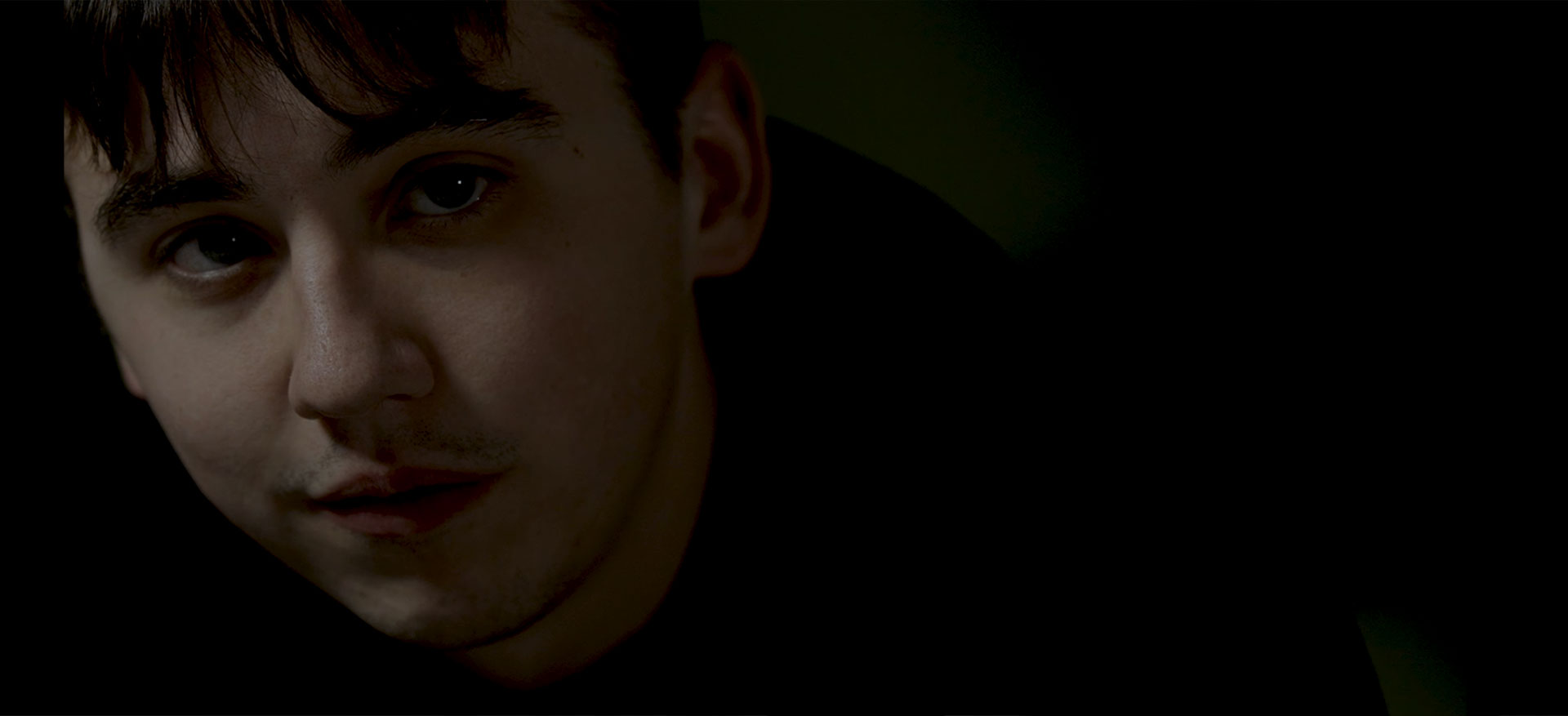 A screenshot from the video showing a boy in a dark room looking somber.